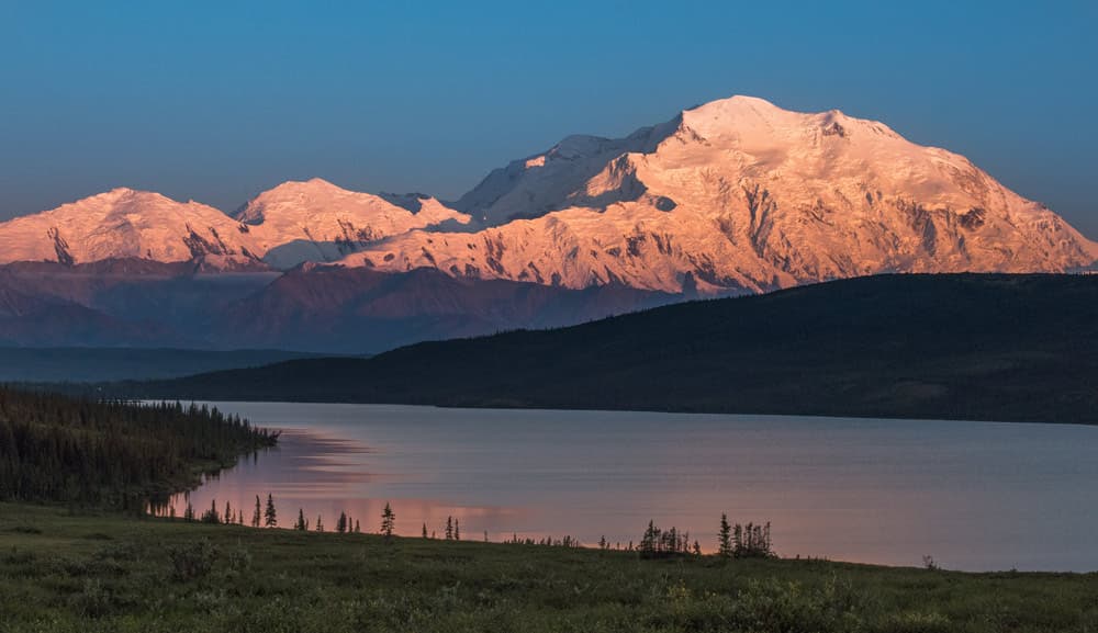 The snow-covered mountain glows pink above dark foothills and a still lake, beneath a blue sky.